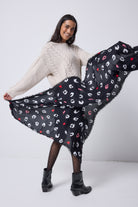 Scamp and Dude Black with White Love Leopard Charity Super Scarf | Model wearing an oatmeal cable knit jumper with a black denim skirt, tights and boots. Holding a black scarf with white love leopard print.