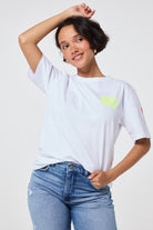 Scamp and Dude White with Neon Yellow Smile Back Slogan T-Shirt | Model wearing white short sleeve crew neck t-shirt which features a neon yellow 'smile back' slogan on the front and back, this is paired with a pair of blue denim jeans.