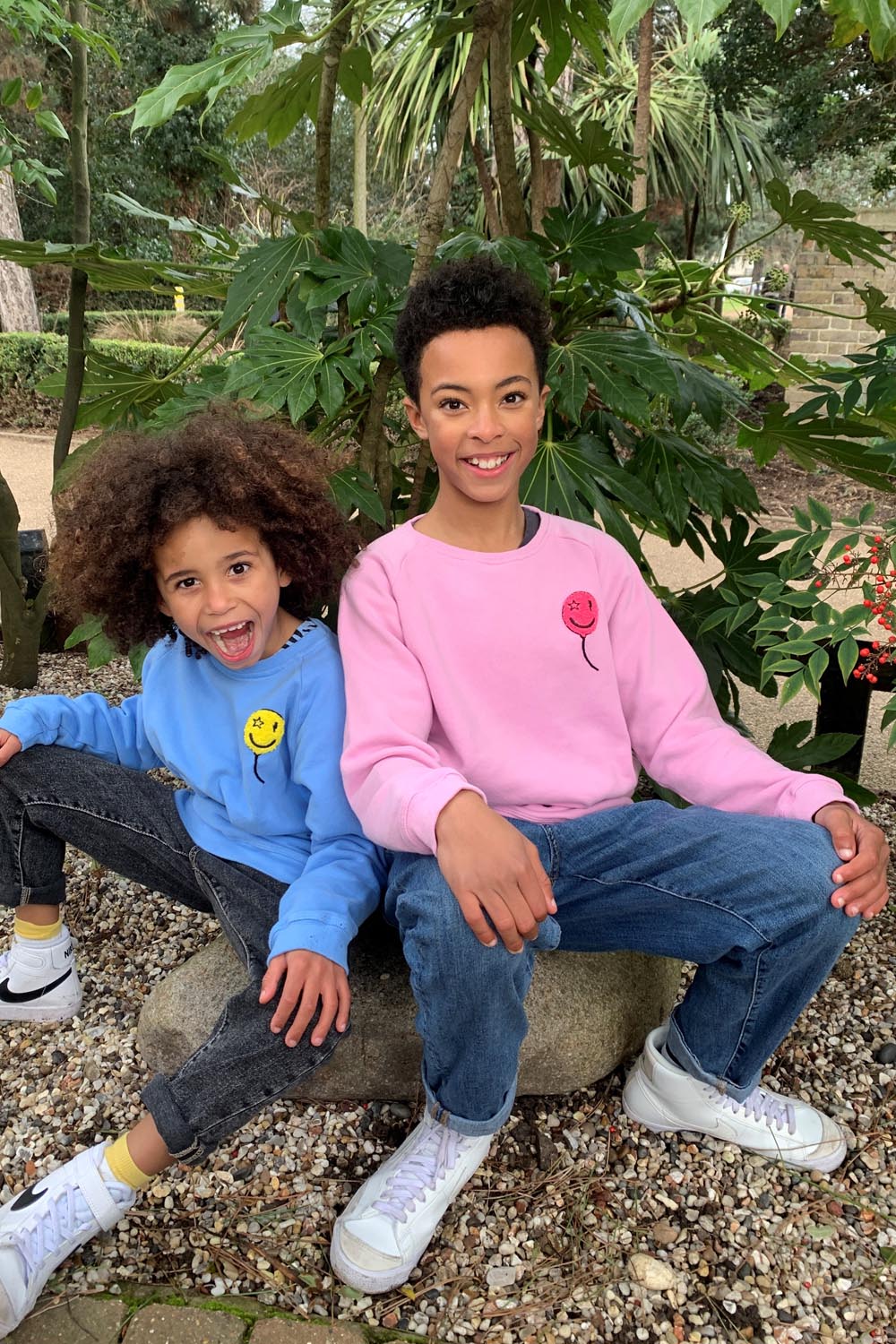 Two boys running in the woods wearing matching sweatshirts in different colourways, one is wearing pink with a neon pink towelling balloon on the chest, the other is wearing blue with a yellow balloon